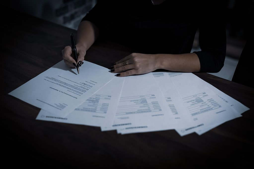 Person signing documents on a dark wood table in dim lighting